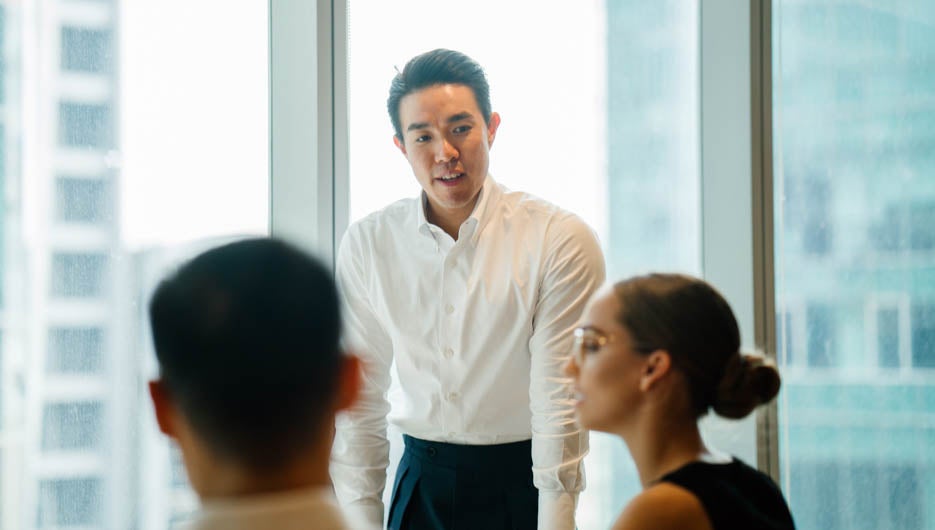 A gorgeous, sure, and fit Asian Chinese man seats a gathering with his group amid the day in the workplace. He is expertly wearing a shirt and pants and is motioning as he talks.