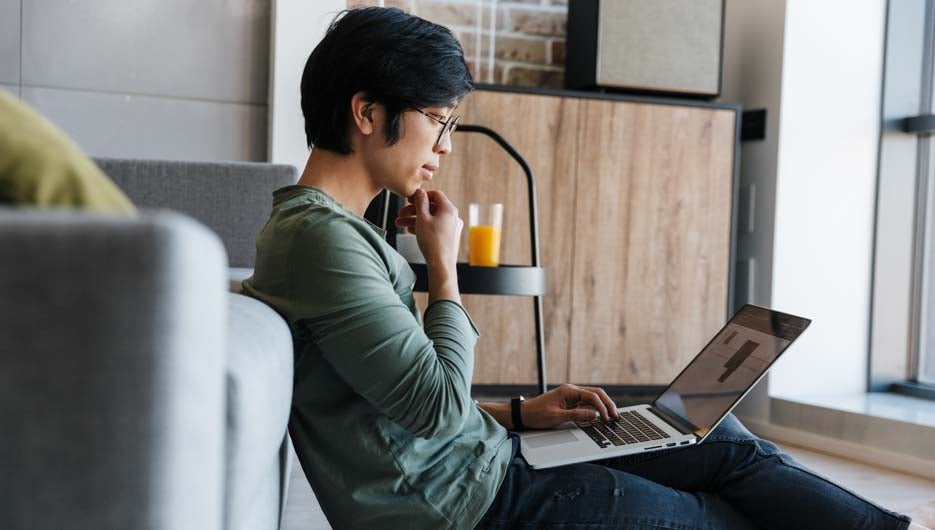 Image of focused asian man in eyeglasses working with laptop while sitting in living room