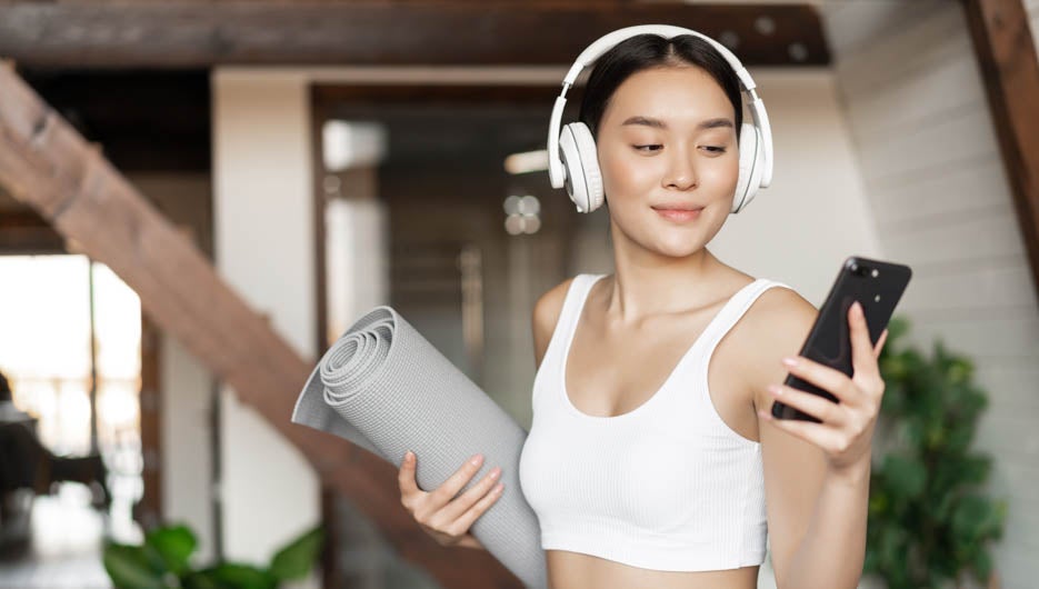 Young asian woman standing at home in headphones, listening music and going workout on floor mat, practice yoga or meditate in living room.