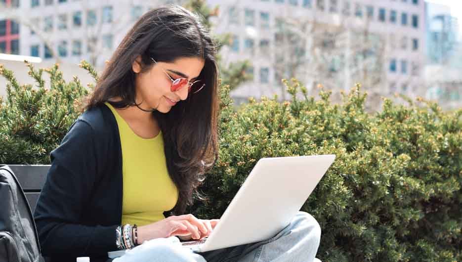 Young happy woman sitting on street bench using laptop computer