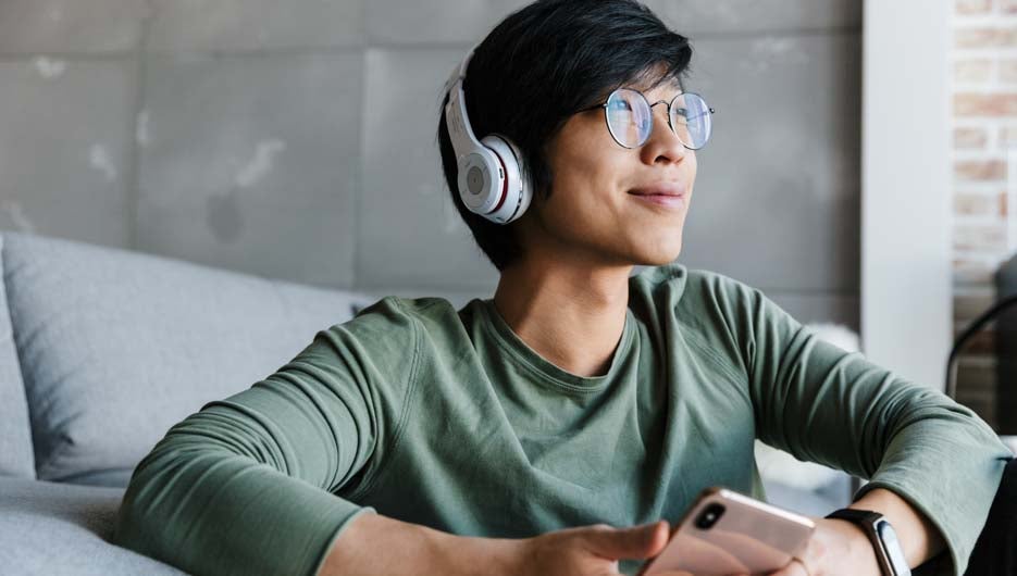 Image of smiling asian man in eyeglasses using cellphone and wireless headphones while sitting in living room
